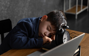 School-age boy sleeping at desk with crooked glasses and small alarm clock nearby. Child smiling...