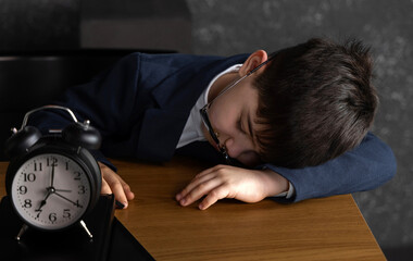 School-age boy sleeping at desk with crooked glasses and small alarm clock nearby. Child smiling...