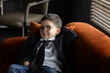 Smiling kid dressed in business jacket with glasses lying on sofa, using laptop and talking on smartphone. Child pretending to be adult, acting like a little boss in office.