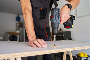 Man in work overalls bib using cordless power drill to make hole in laminated wood board. Male carpenter working in home workshop, drilling chipboard on workbench