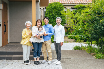 住宅の前で笑顔で赤ちゃんを囲む三世代家族