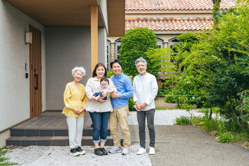 A three-generation family smiling and surrounding a baby in front of their house