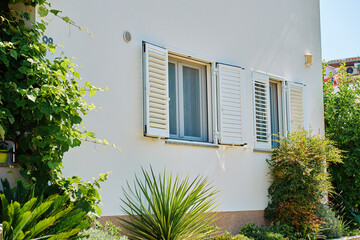 White suburban house wall with two windows, open shutters and plants in foreground. Residential...
