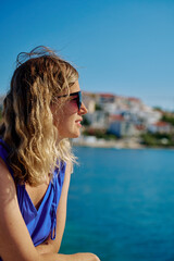 Naklejka premium Side view of woman in sunglasses and blue dress looking at the sea on sunny day. Female tourist relaxing by sea. Concept of relaxation, travel and summer vacation