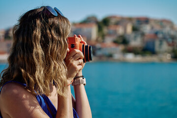 Woman holding orange instant camera, taking photo near sea with blurred town in background. Female...