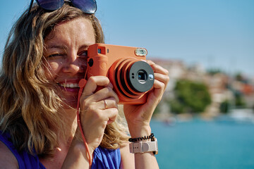 Woman standing near sea holding instant camera and taking photo. Female tourist at coastal resort...