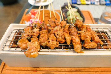 Freshly grilled chicken satay skewers served on a small tabletop grill glazed and slightly charred, served with peanut sauce and traditional condiments in the background. Ideal for culinary content
