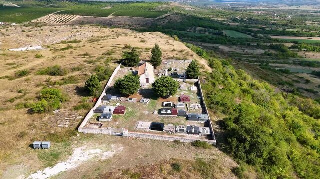 Aerial view of a rural cemetery in Asseria, Croatia with scattered tombstones and a small chapel, surrounded by a stone wall and trees, set in a vast landscape with fields and distant hills.