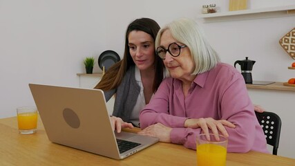 Young woman teaching her grandmother how to use laptop computer sitting together in the kitchen at home, enjoying quality time and connection in their home while drinking homemade orange juice - Powered by Adobe
