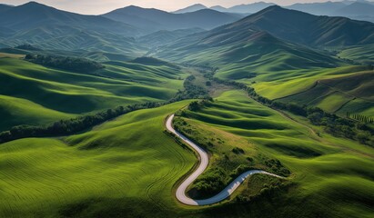 Aerial view of lush green rolling hills and winding road through natural landscape under blue sky with morning sunlight, showcasing springtime beauty of grassy fields and distant mountains