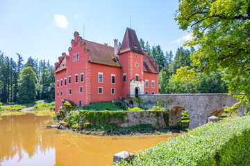 Obraz premium Cervena Lhota Castle is situated on a rocky island at the center of a lake, featuring vibrant red walls and historical architecture amidst a lush green landscape in Czechia.