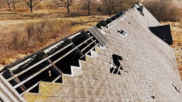 Drone glides over shattered asbestos roof of Soviet-era barn, rural spring field