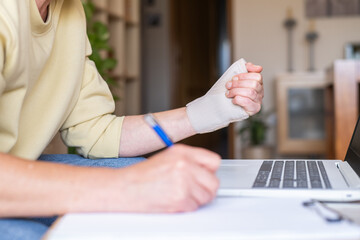Woman with rhizarthrosis and tendonitis using a laptop and taking notes at home, her injured hand...