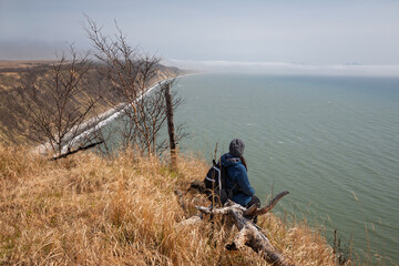 A woman sits on a dead tree trunk on a high seashore and looks into the distance at the sea. Travel and tourism in the Magadan region. The coast of the Sea of ​​Okhotsk. Nature of the Russian Far East