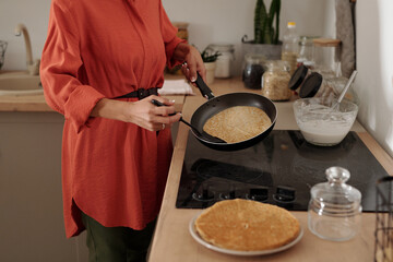 Unrecognizable woman preparing pancakes in kitchen, holding frying pan and spatula, standing near stove with stack of pancakes, focusing on cooking process