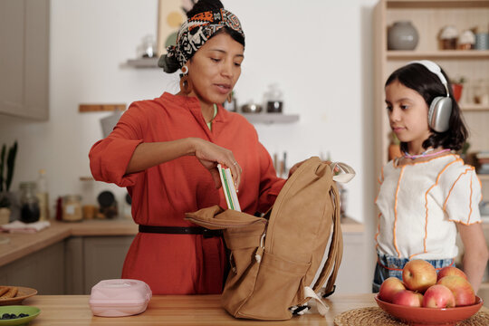 Hispanic woman packing books into backpack while her daughter wearing headphones standing nearby in kitchen preparing for school - Powered by Adobe