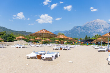 Tekirova, Turkey - June 12, 2025: view of an organized beach area with rows of empty sun loungers and straw umbrellas. Mountains and forest visible in the background under a bright blue sky. Perfect
