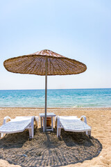 Two white sun loungers under a straw parasol on a sandy beach with turquoise sea and clear blue sky. Idyllic summer vacation concept.