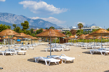 Tekirova, Turkey - June 12, 2025: view of an organized beach area with rows of empty sun loungers and straw umbrellas. Mountains and forest visible in the background under a bright blue sky. Perfect