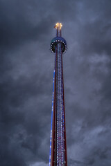 A striking and breathtaking view of an illuminated amusement park tower piercing through a dramatic and moody cloudy sky, showcasing vibrant colors and dazzling lights, Szczecin, Poland