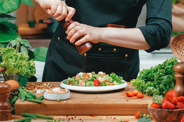 Chef skillfully preparing afresh salad in kitchen. Using pepper mill, add seasoning to plate with crisp lettuce, strawberries and garnishes. Various fresh ingredients, herbs and vegetables