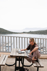 Fototapeta premium Young woman working remotely by the sea with a laptop and solar panel, embracing sustainable travel, mindful lifestyle and digital freedom. Concept of new work, balance and conscious living.
