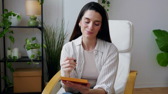Mid adult woman sitting comfortably in armchair, writing personal notes with focus and concentration, deeply immersed in thoughtful creative process while working from home environment