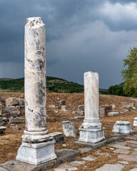 Ancient weathered marble columns stand among ruins at the Asklepion Ruins in Bergama, Turkey, archaeological site under a dramatic stormy sky. Historical landscape