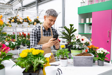 A smiling man in an apron uses his smartphone to take a photo of plants in a greenhouse.
