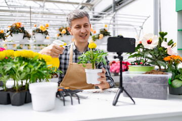 A man, wearing an apron, shows a potted flower while live streaming from a greenhouse.