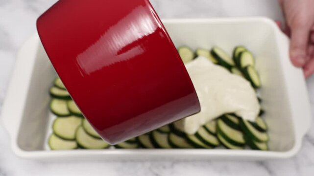 Chef is using a spoon to spread bechamel sauce over sliced zucchini arranged in a white baking dish, preparing a delicious and healthy vegetarian meal