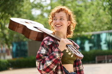 Young musician carrying acoustic guitar in a park looking away smiling