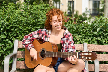 Young redhead woman playing acoustic guitar on park bench