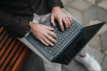 Close-up view of hands typing on a laptop held by a person wearing casual attire, seated on a wooden bench in an outdoor setting. Focus on technology and casual work environment.