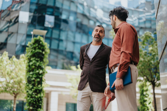 Two coworkers stand outside an office building, engaging in a serious conversation while holding business materials. This urban setting is enhanced by modern architecture and green surroundings. - Powered by Adobe