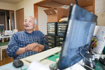 In Japan, inside a small apartment room, a Japanese man in his late 70s sits in front of a computer, smiling and chatting, with warm indoor lighting creating a peaceful and homely atmosphere.