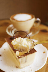 Closeup of a Spoon Scooping Mouthwatering Tiramisu with Blurry Cappuccino in the Backdrop