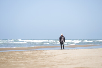 By the rippling Sea of Japan on a sunny May day, a Japanese man in his late 70s wearing a checkered shirt stands alone on the beach. Gentle waves and spring air evoke a quiet moment of reflection.