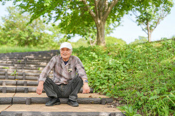 Fototapeta premium In a lush green park on a sunny May day, a Japanese man in his late 70s, wearing a checkered shirt and white hat, sits on the steps. Surrounded by spring foliage, he enjoys a quiet, peaceful moment.