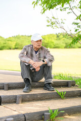 Fototapeta premium In a lush green park on a sunny May day, a Japanese man in his late 70s, wearing a checkered shirt and white hat, sits on the steps. Surrounded by spring foliage, he enjoys a quiet, peaceful moment.