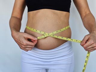 Overweight and obese woman in sports bra holding tape measure around fat belly to check how much...