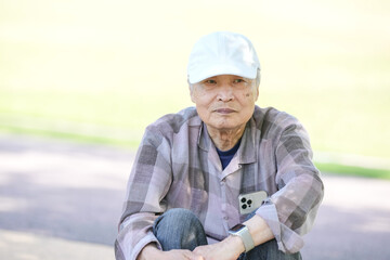 In a lush green park on a sunny May day, a Japanese man in his late 70s, wearing a checkered shirt and white hat, sits on the steps. Surrounded by spring foliage, he enjoys a quiet, peaceful moment.
