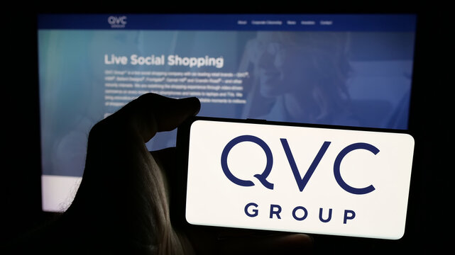 Stuttgart, Germany - 06-07-2025: Person holding mobile phone with logo of US media company QVC Group Inc. on screen in front of web page. Focus on phone display.