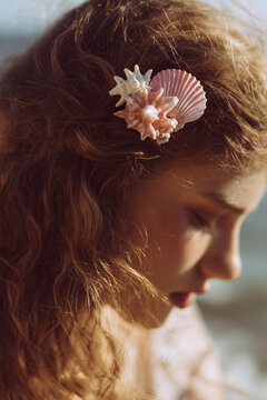 A dreamy close-up of a woman's hair styled with a delicate pink-and-white sea shell hair clip, shaped like coral or a spiked conch. The soft natural light creates a nostalgic, coastal feel. 