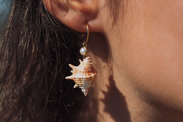 Close-up of a sea shell earring with pearl on ear
A detailed view of an elegant earring made from a real spiral sea shell, delicately hanging from a woman's ear. 