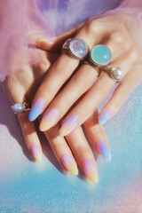 Aura-inspired nail art with bold statement rings on pastel background
A close-up of hands with almond-shaped nails, painted in soft ombré gradients resembling an aura or cosmic energy. 