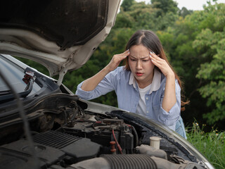 A young woman in a jeans shirt with a broken car with the hood open holds her head in her hands.