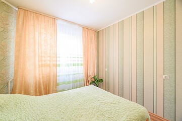 cozy bedroom with a green fluffy bedspread, peach curtains by the window, and striped wallpaper
