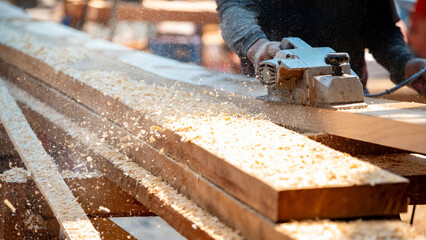 Male carpenter hand using an electric planer on wooden plank at construction site. Woodworker planing timber in his workshop with sawdust spreading around. Lumber industry and carpentry work tool