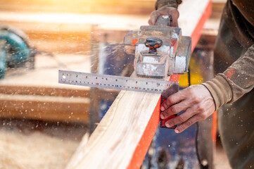 Male carpenter hand using angle ruler for wood squaring and planing on wooden plank by electric planer on workbench in workshop. Wood measuring and shaving process for lumber industry